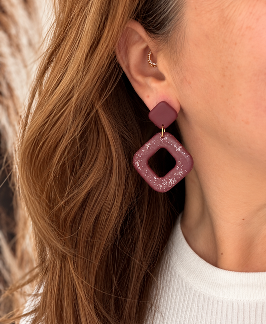 Burgundy statement oorbellen met een vierkante stud en een open, ruitvormige hanger met glinsterende glitterdeeltjes en een goudkleurig verbindingsringetje.